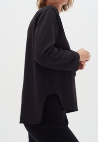 Maglietta nera a maniche lunghe con scollatura rotonda, vestibilità rilassata e orlo asimmetrico. Tessuto morbido con texture minimale, abbinata a un pantalone nero.
