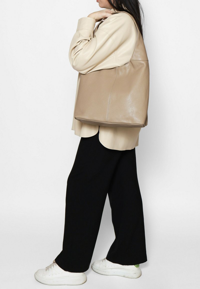 Beige leren tote bag met een gladde textuur, ruime ontwerp en geen zichtbare hardware, gedragen over een beige shirt en zwarte broek.