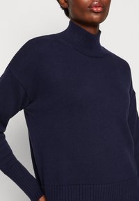 Maglione a collo alto blu navy realizzato in tessuto a maglia testurizzata, con polsini e orlo a coste, vestibilità comoda e fessure laterali per facilitare il movimento.