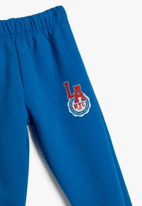 Niebieskie dresy z elastycznym paskiem w talii, z czerwoną i białą grafiką "LA NYC" oraz okrągłym logo po prawej nogawce. Materiał bawełniany.