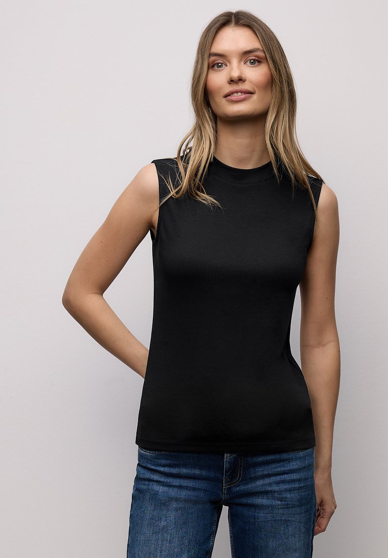 Femme aux cheveux blonds longs portant un haut noir sans manches et un jean bleu, debout avec une main derrière le dos contre un mur uni.