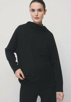 Top negro de manga larga con cuello tipo embudo, tela suave, corte relajado y aberturas laterales. Combinado con pantalones negros a juego.