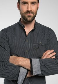 Gestreepte overhemdbloes in zwart en wit met een borstzak, featuring een hertembleem. Grijze manchetten met zwarte accenten. Zachte stoftextuur.