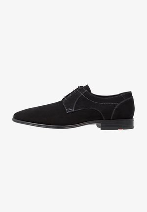 Chaussure noire en suède à lacets avec un profil bas, présentant des coutures contrastantes et une semelle lisse. Design simple avec un bout arrondi.
