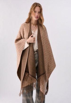Donna con capelli lunghi che indossa uno scialle beige a motivo sopra una camicetta bianca e pantaloni a quadri con collane d'oro sovrapposte e cintura nera.