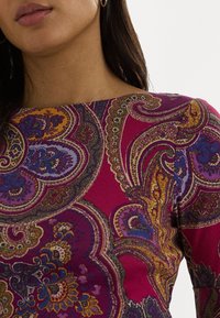 Mujer que lleva una blusa de manga larga con vibrantes patrones de paisley en púrpura, amarillo y azul, y pendientes de aro dorados, con el cabello castaño oscuro a la vista.
