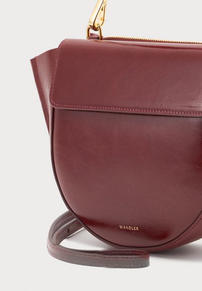 Σκούρο κόκκινο δερμάτινο τσαντάκι crossbody με κλείσιμο καπακιού, χρυστό φερμουάρ και χρυσό κούμπωμα, με καμπυλωτό κάτω μέρος και ιμάντα που είναι τυλιγμένος στη βάση.