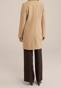 Beige villakangastakki, jossa suora siluetti ja lovikaulus, yhdistetty ruskeisiin leveälahkeisiin housuihin ja tummiin korkokenkiin.