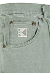 Jeans en denim vert clair avec un logo cousu sur la poche arrière, des rivets en bronze et une finition en tissu texturé.