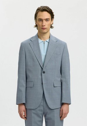 Junger Mann mit mittellangen Haaren, der eine hellgraue Anzugsjacke und passende Hose über einem hellblauen Reißverschlusshemd trägt.