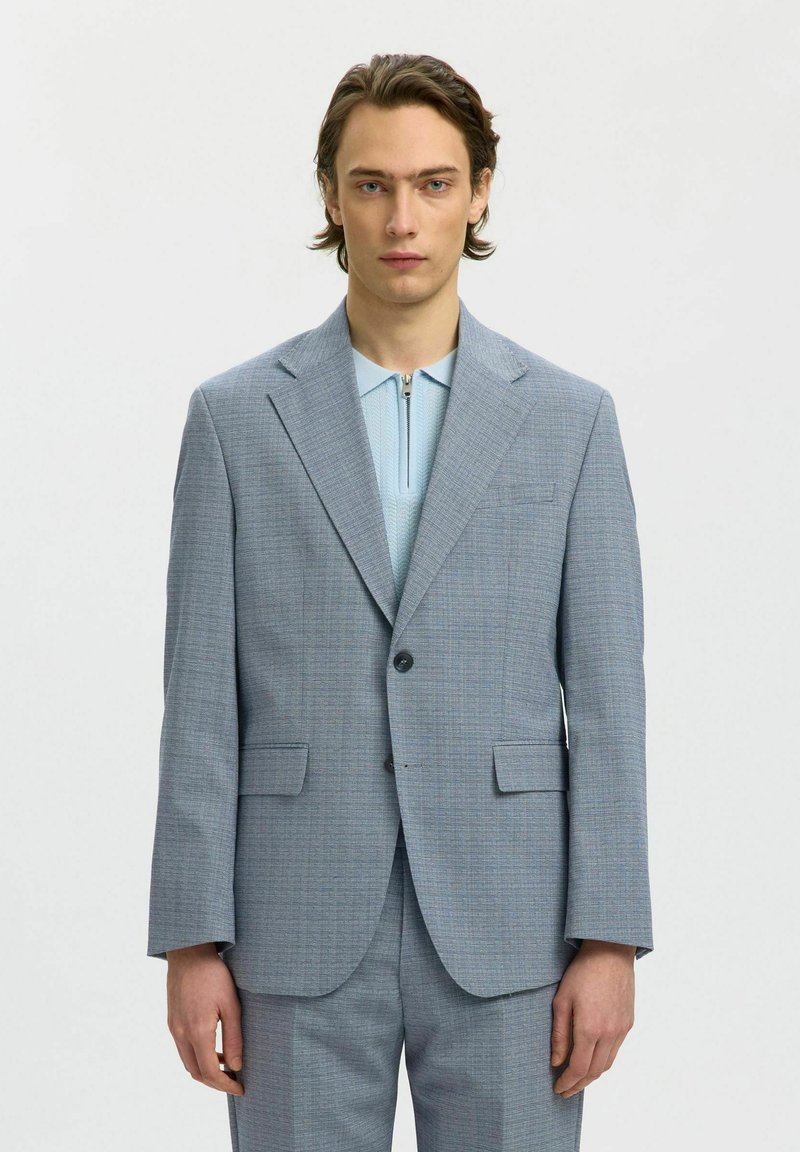 Jeune homme aux cheveux mi-longs portant une veste de costume gris clair et un pantalon assorti par-dessus une chemise bleu clair zippée.