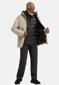 Veste imperméable beige par-dessus un gilet matelassé noir, tous deux avec des fermetures éclair. Un pantalon sombre et des baskets noires complètent la tenue.