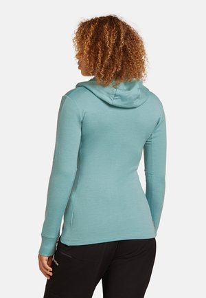 Kringelhaarige persoon in een aansluitende teal kleurige hoodie met lange mouwen en zwarte broek, met de rug naar de kijker gekeerd.