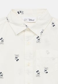 Πουκάμισο με κοντά μανίκια σε εκρού βαμβάκι με μαύρα σχέδια του Μίκυ Μάους. Γιακάς με πτύχωση, κουμπιά μπροστά και τσέπη στο στήθος με σχέδιο Μίκυ.