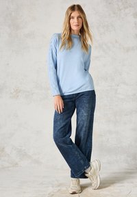 Hellblaue Langarmbluse mit strukturierten Details an den Ärmeln, kombiniert mit dunkelblauen weiten Jeans und weißen Sneakers, vor einem neutralen Hintergrund.