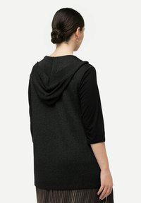 Gilet nero senza maniche con cappuccio, tessuto testurizzato, taglio dritto e una leggera lucentezza. Indossato sopra un top nero aderente e una gonna plissettata.