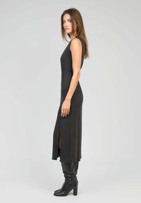 Robe maxi noire sans manches avec une fente sur le côté, fabriquée dans un tissu lisse. Associée à des bottes noires hautes et ajustées. Design simple et épuré.
