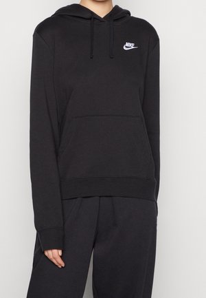 Personne portant un sweat à capuche Nike noir uni avec un logo blanc sur la poitrine et un pantalon noir assorti sur fond uni.