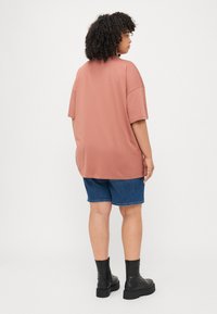 Camiseta marrón de gran tamaño, de mangas cortas, combinada con pantalones cortos de denim azul. Presenta un corte relajado, una textura suave y botas negras hasta el tobillo.