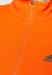 Oranssi vetoketjullinen takki, joka on valmistettu pehmeästä kankaasta korkean kauluksen kanssa. Takissa on musta Adidas-logo ja siihen sopiva vetoketju. Sujuva rakenne, urheilullinen muotoilu.