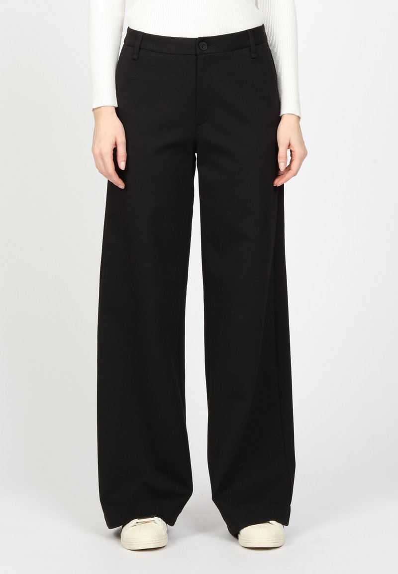 Femme portant un pantalon large noir, un haut à manches longues blanc à côtes et des baskets blanches, se tenant devant un fond uni.