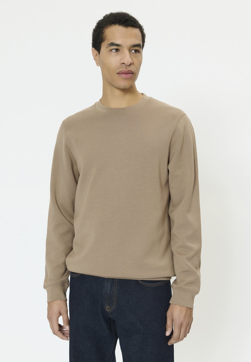 Mees, kes kannab beežikat pikkade varrukatega särki ja tumedaid teksapükse, seisab tavalise valge tausta ees ning vaatab kergelt küljele.