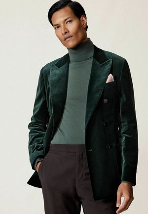 Tmavozelený zamatový blazer s špicatými klopami, jednoradovým strihom a čiernymi gombíkmi. Nosený cez zelený rolák.
