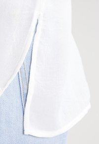 White, flowing linen top with a rounded hem, featuring a raw edge. Paired with light blue denim, showing a side seam and stitching detail.