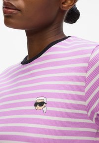 Lila- und weiße gestreifte Bluse aus geripptem Stoff mit einem schwarzen Halsauschnitt, die ein kleines gesticktes Maskottchen mit Sonnenbrille zeigt.