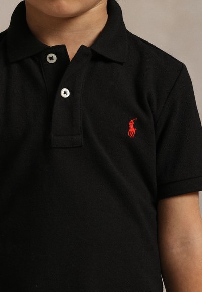 Dieťa má na sebe čierne polo tričko s krátkym rukávom, bielymi gombíkmi a malým červeným vyšívaným logom hráča póla na hrudi.
