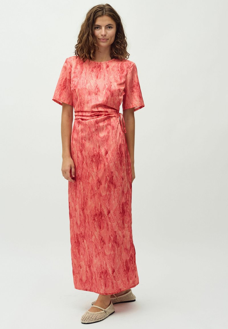 Robe longue rose avec un motif texturé, manches courtes et une ceinture nouée à la taille. Associée à des chaussures à plat neutres et à motifs.