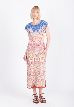 Ženska z blond bob pričesko, oblečena v barvito vzorčasto midi obleko in rjave sandale z naramnicami, stoji pred belim ozadjem.