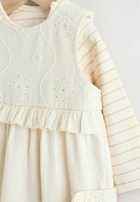 Robe en coton blanc avec broderies florales, ourlet volanté, superposée à un haut à manches longues rayé crème et rose. Texture douce, design décontracté.
