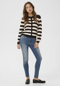 Melna un bežā svītru jaka ar pogām, apvienota ar pieguļošām zilām džinsām un melniem loafers. Dizains ir ikdienišķs un mūsdienīgs.