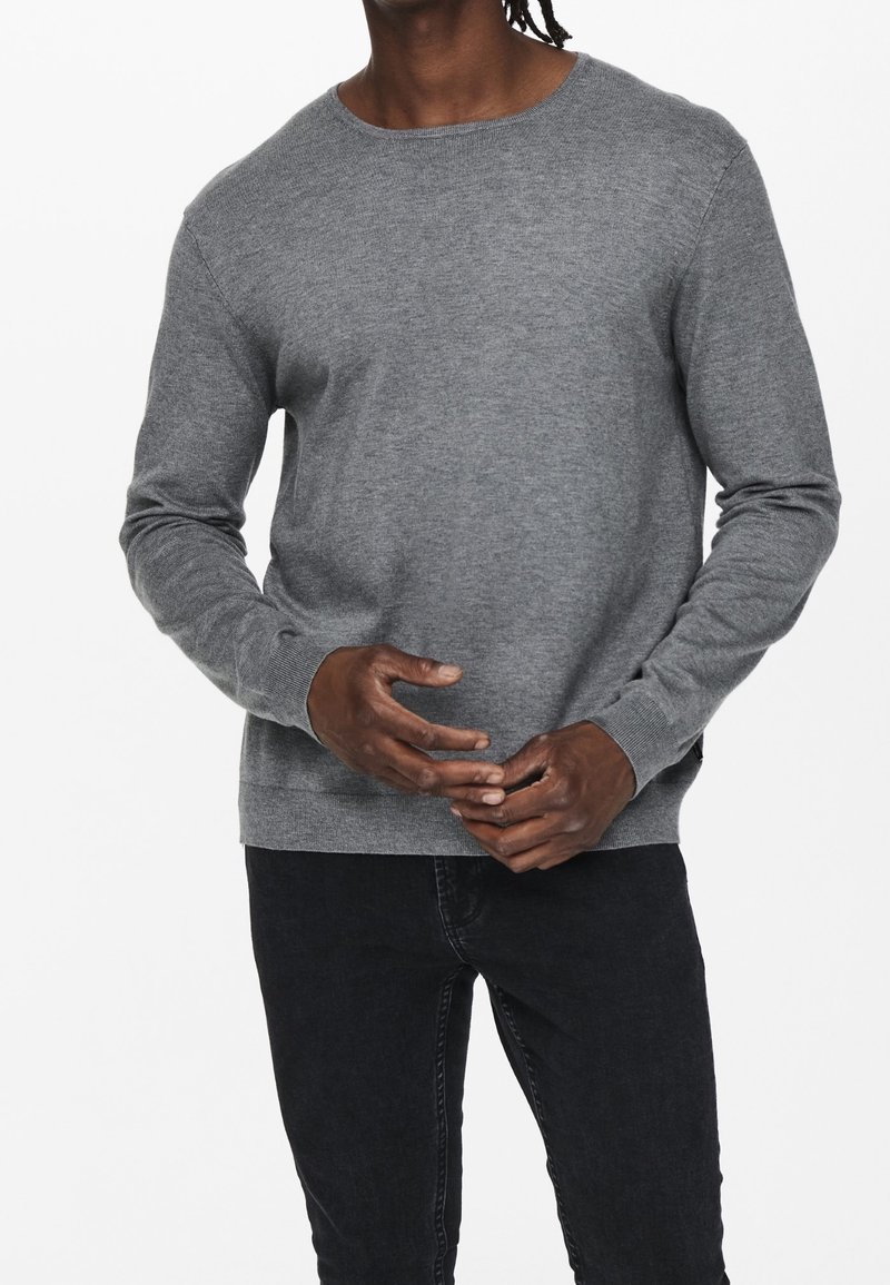 Homme portant un pull gris uni à manches longues et un jean noir, debout devant un fond blanc, les mains jointes devant lui.