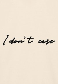 T-shirt beige con testo nero scritto a mano "Non mi interessa" al centro. Tessuto morbido, collo a giro standard, vestibilità casual.