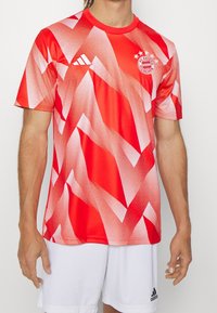 Camiseta deportiva roja de manga corta con patrones abstractos en blanco, que lleva el logo de Adidas y el emblema del FC Bayern Munich en el pecho.