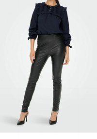 Blouse marine à design plissé et détails de boutons, associée à un pantalon noir en simili cuir taille haute et à des talons hauts noirs.