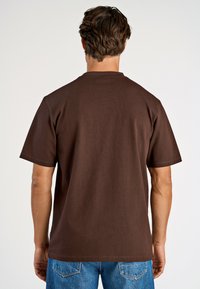 Brun bomulls t-shirt med korta ärmar och en avslappnad passform. Har rundad halsringning och en mjuk textur. Bärs med blå denimjeans.