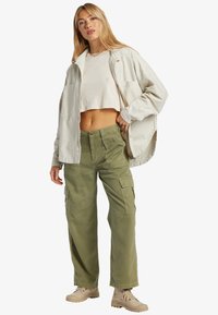 Light cream cropped jacket over a white cropped shirt paired with loose, olive green cargo pants featuring pockets and a button closure.