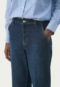 Jeans de denim azul oscuro con corte recto, con costuras doradas, bolsillos delanteros y cierre de botón. Combinados con una camisa a rayas azules.