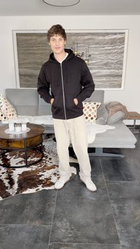 Haut de hoodie zippé noir avec poches avant, pantalon beige clair et baskets blanches. L'arrière-plan présente un canapé gris et une table ronde en bois.