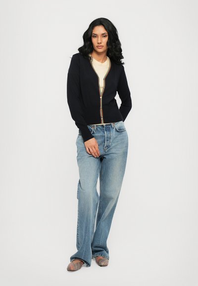 Cardigan navy con scollo a V, rifiniture colorate a contrasto e chiusura con bottoni, abbinato a jeans azzurri e scarpe a righe multicolori.