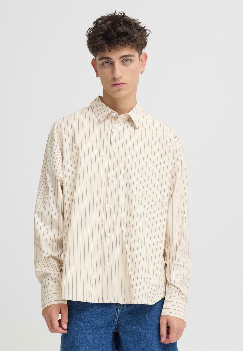 Chemise beige à boutons avec rayures verticales bleues, manches longues et une poche poitrine. Le tissu semble lisse avec une coupe décontractée.