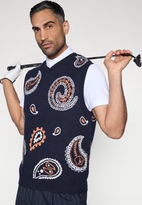 Tamsiai mėlynas megztas golfo liemenėlis su paisley raštais baltos ir oranžinės spalvos, V formos iškirpte ir raukšlėtomis rankovių angomis. Golfo žaidėjas laiko klubą.