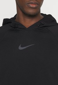 Muž v čierneho štruktúrovaného Nike mikiny s kapucňou a nastaviteľnými šnúrkami, na hrudi má šedý Nike swoosh logo.