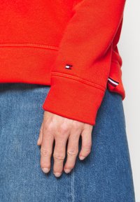 Felpa rossa con polsini a costine, dotata di un piccolo logo del marchio sulla manica. Indossata con jeans in denim azzurro chiaro.