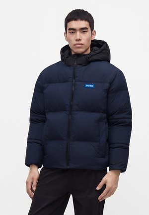 Jeune homme portant une doudoune bleu marine avec une capuche noire et un patch logo Hugo bleu sur la poitrine, debout devant un fond uni.
