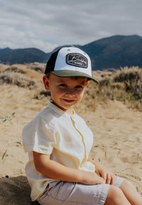 Gorra de béisbol con parte frontal blanca, lados negros y visera verde; cuenta con un parche de logo. El niño lleva una camisa de color amarillo claro con cuello.
