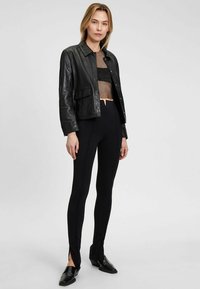 Giubbotto di pelle nera con colletto, indossato sopra un top in rete corto. Leggings neri a vita alta con spacchi laterali e texture a coste.